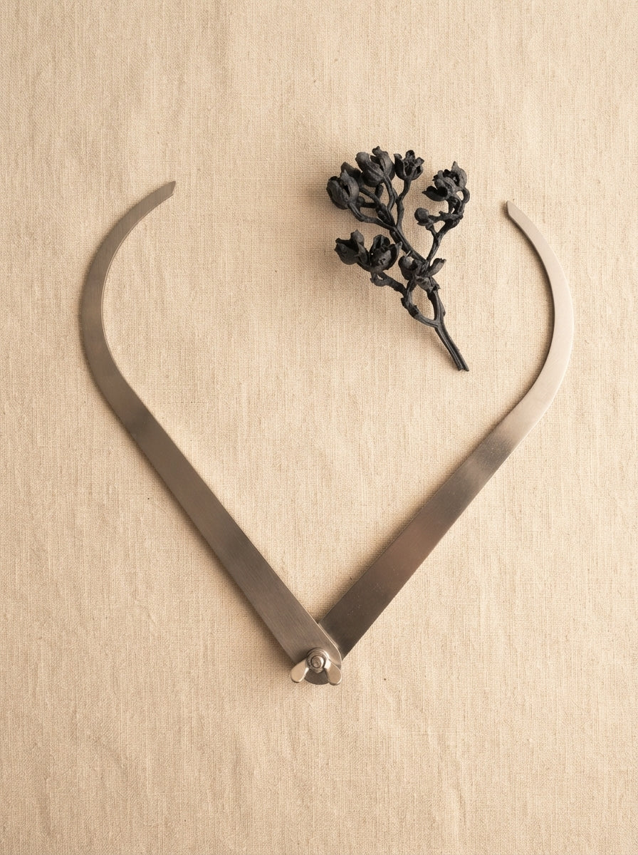 Stainless steel 10-inch clay caliper laid flat on a warm cream linen surface with a small dried charcoal plant sprig.